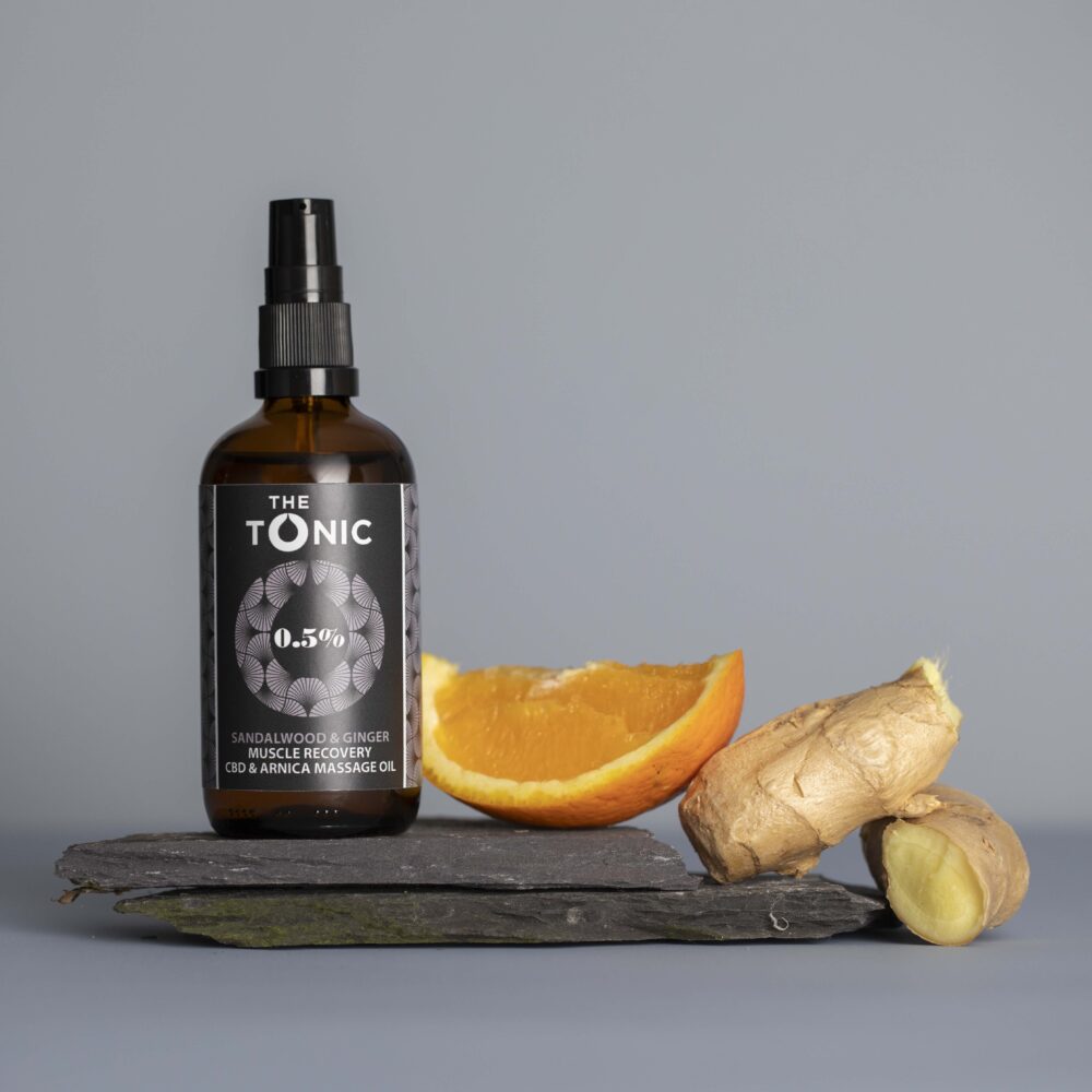 Muscle Recovery Massage oil bottle stands upon a piece of rustic slate, with a slice of orange and pieces of fresh ginger to depict the essential oils added to this massage oil, alongside arnica and CBD.